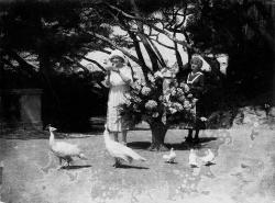 Dans un jardin, devant une grosse corbeille de fleurs, une femme lit une lettre en présence d'une domestique : Gaby Deslys