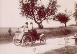 C’est l’une de ces premières voitures qui est ici photographiée sur une route de campagne. Le chauffeur et son passager sont vus de dos, et l’on distingue le moteur placé à l’arrière.