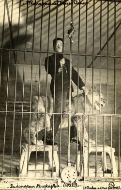 en équilibre sur deux tabourets de bois, le dompteur chevauche le jeune lion qu’il tient fermement par son collier.